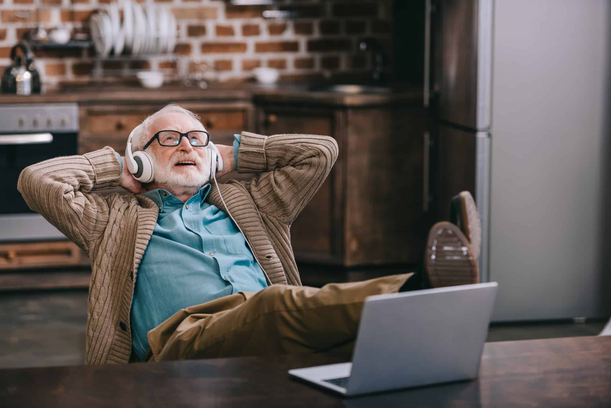 Happy old man in headphones using laptop with feet on table